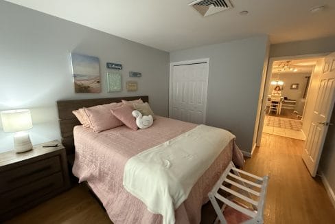 Bedroom with a queen bed, pink bedding, two bedside tables, a lamp, wall art above the headboard, a folded towel on the bed, and a view into a hallway with dining area.