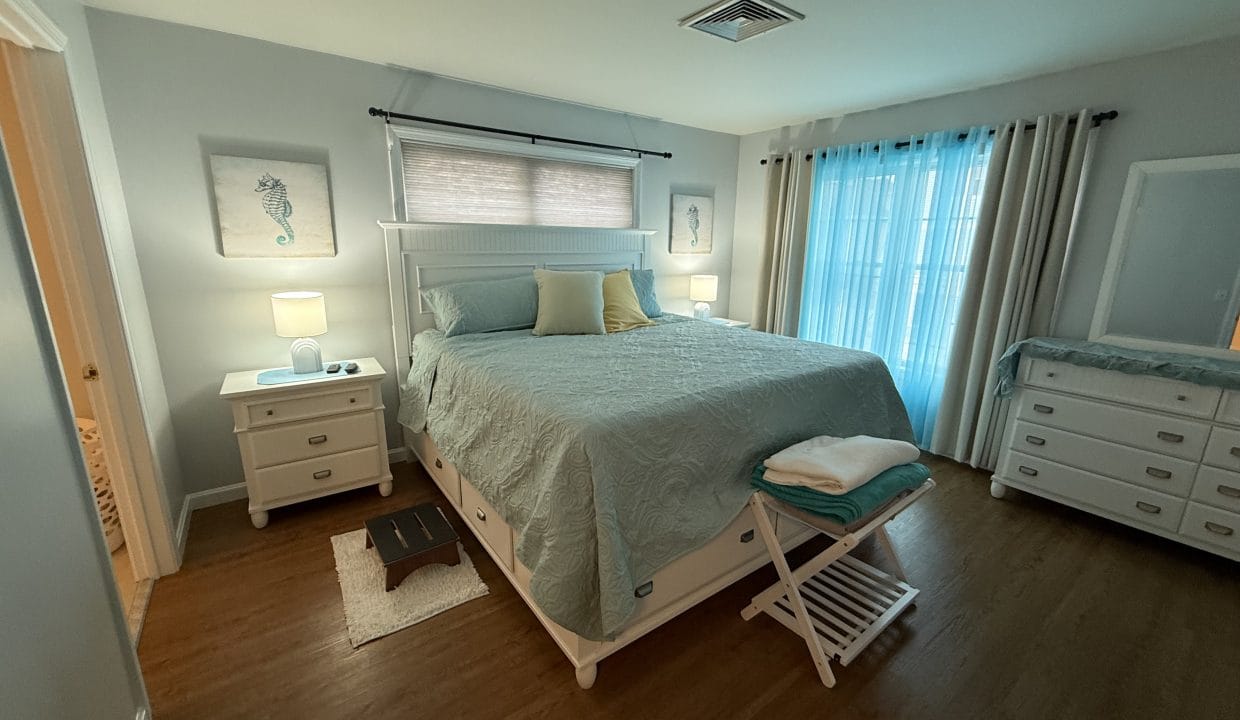 A neatly made bed with seafoam bedding is centered in a bright bedroom with two nightstands, a dresser, artwork, and sheer curtains over a window.