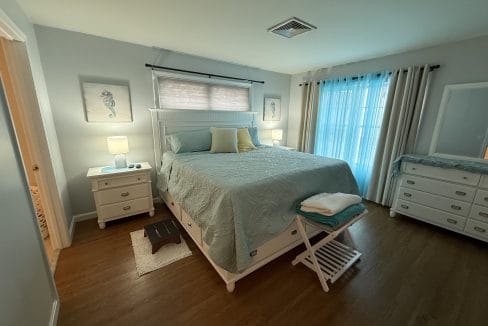 A neatly made bed with seafoam bedding is centered in a bright bedroom with two nightstands, a dresser, artwork, and sheer curtains over a window.
