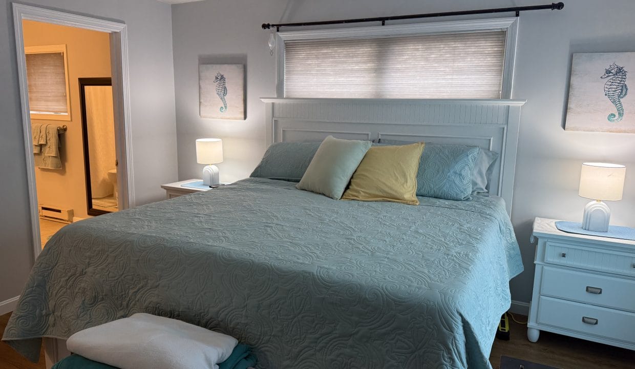 A neatly made bed with blue and yellow pillows, two nightstands with lamps, seahorse art on the wall, and an adjacent bathroom visible.