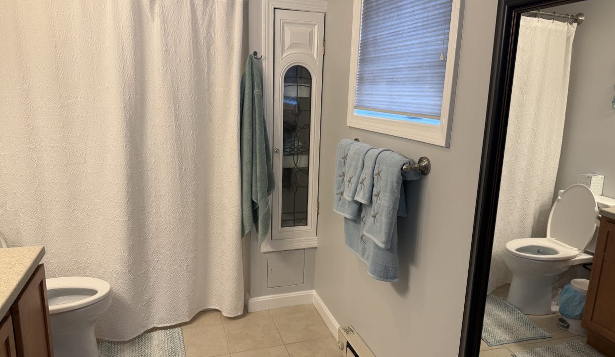 A bathroom with a toilet, shower with white curtain, vanity, wall-mounted towels, mirror, window with a shade, and beige tiled floor.
