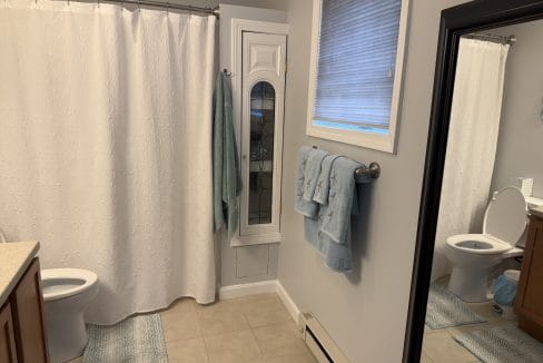 A bathroom with a toilet, shower with white curtain, vanity, wall-mounted towels, mirror, window with a shade, and beige tiled floor.