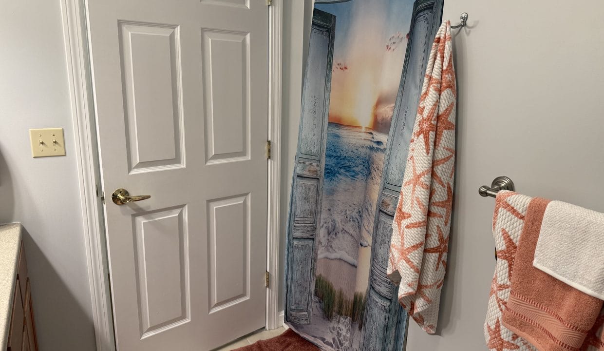 A bathroom with a white door, ocean-themed shower curtain, white and coral towels with starfish patterns, and a brown bath mat on a tiled floor.