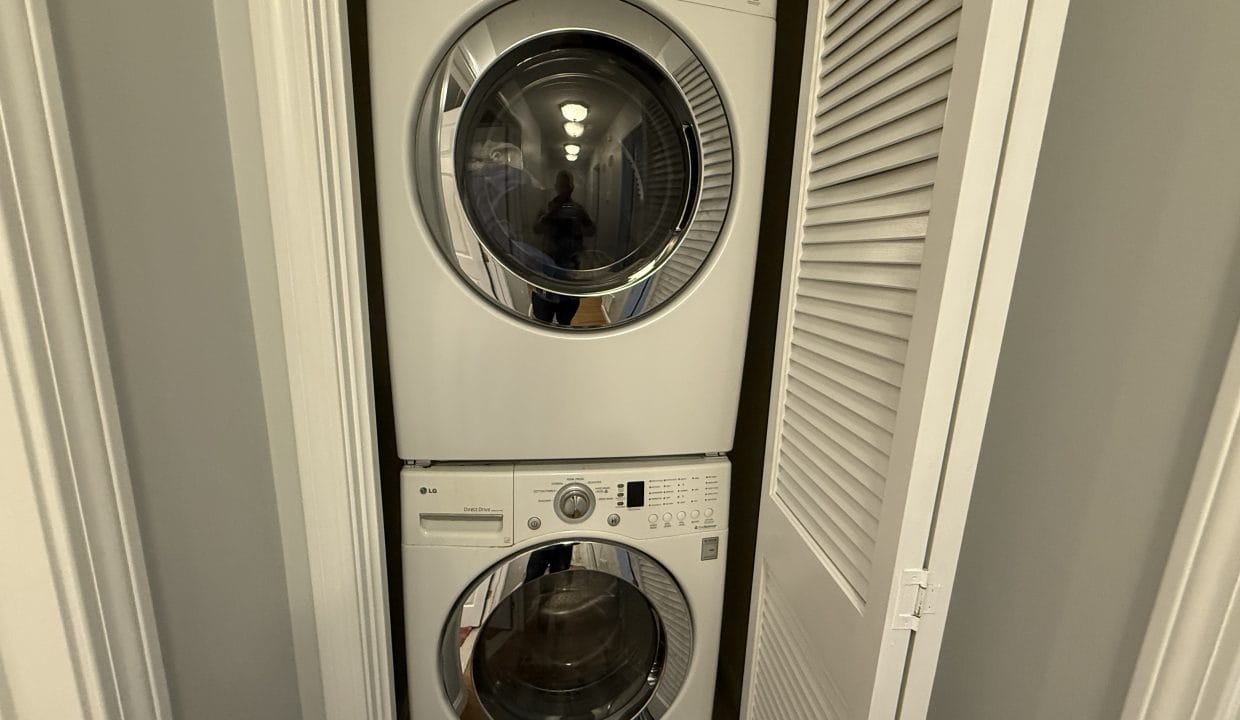 Stacked front-loading washer and dryer units in a small closet with white louvered doors partially open.