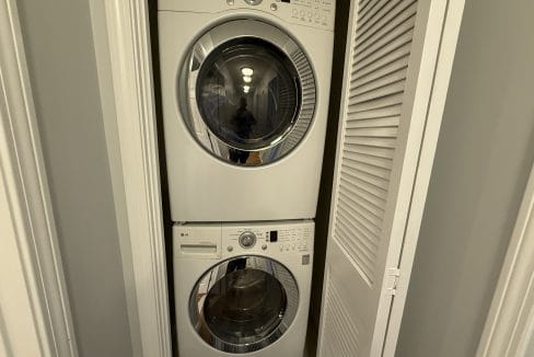 Stacked front-loading washer and dryer units in a small closet with white louvered doors partially open.