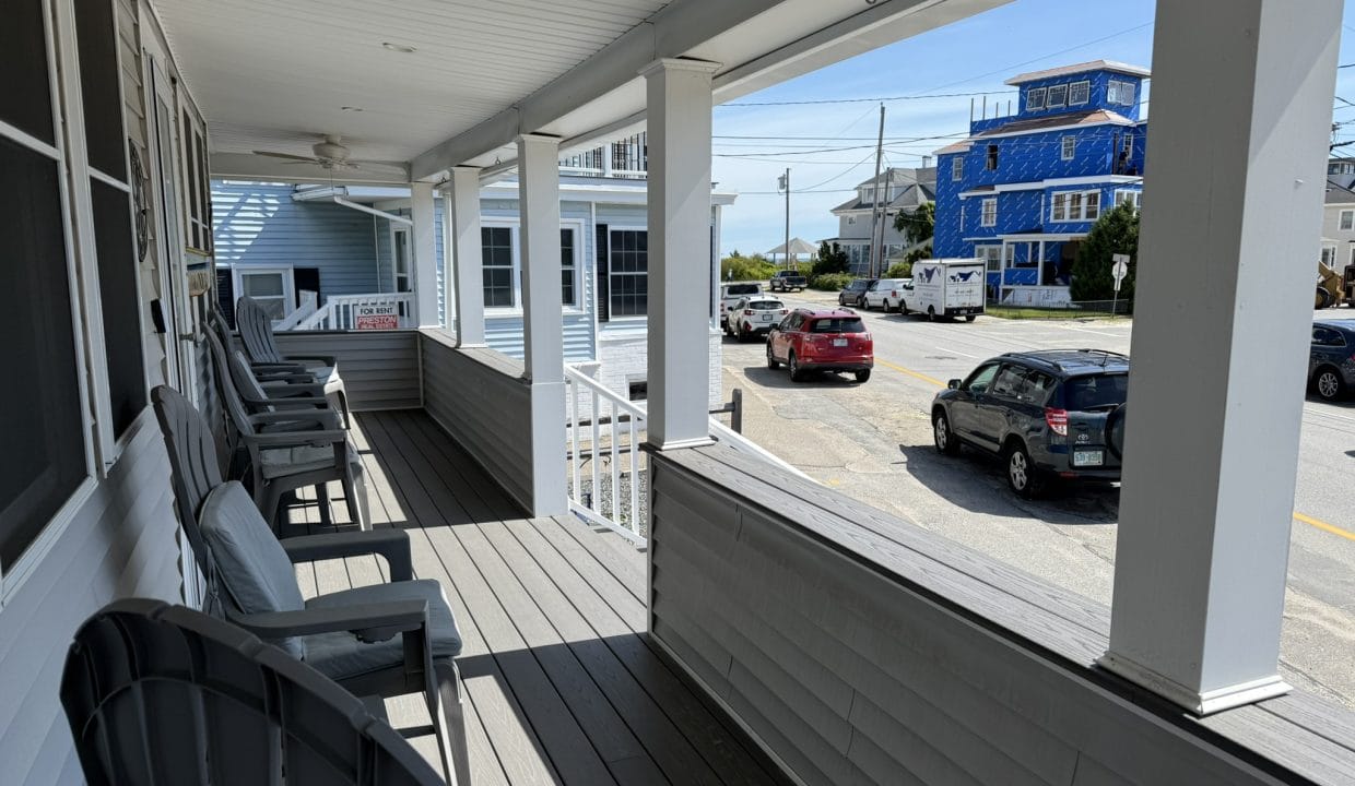 A covered porch with Adirondack chairs overlooks a residential street with parked cars and houses, including a blue house under construction.