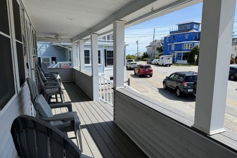 A covered porch with Adirondack chairs overlooks a residential street with parked cars and houses, including a blue house under construction.