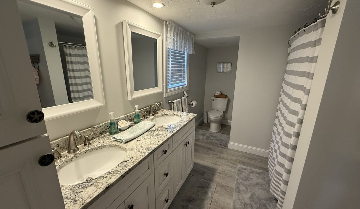 Bathroom with double sink vanity, granite countertop, mirror, striped shower curtain, window, toilet, and gray floor mats on light wood flooring.