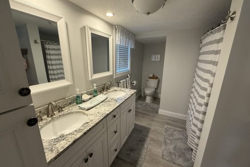 Bathroom with double sink vanity, granite countertop, mirror, striped shower curtain, window, toilet, and gray floor mats on light wood flooring.
