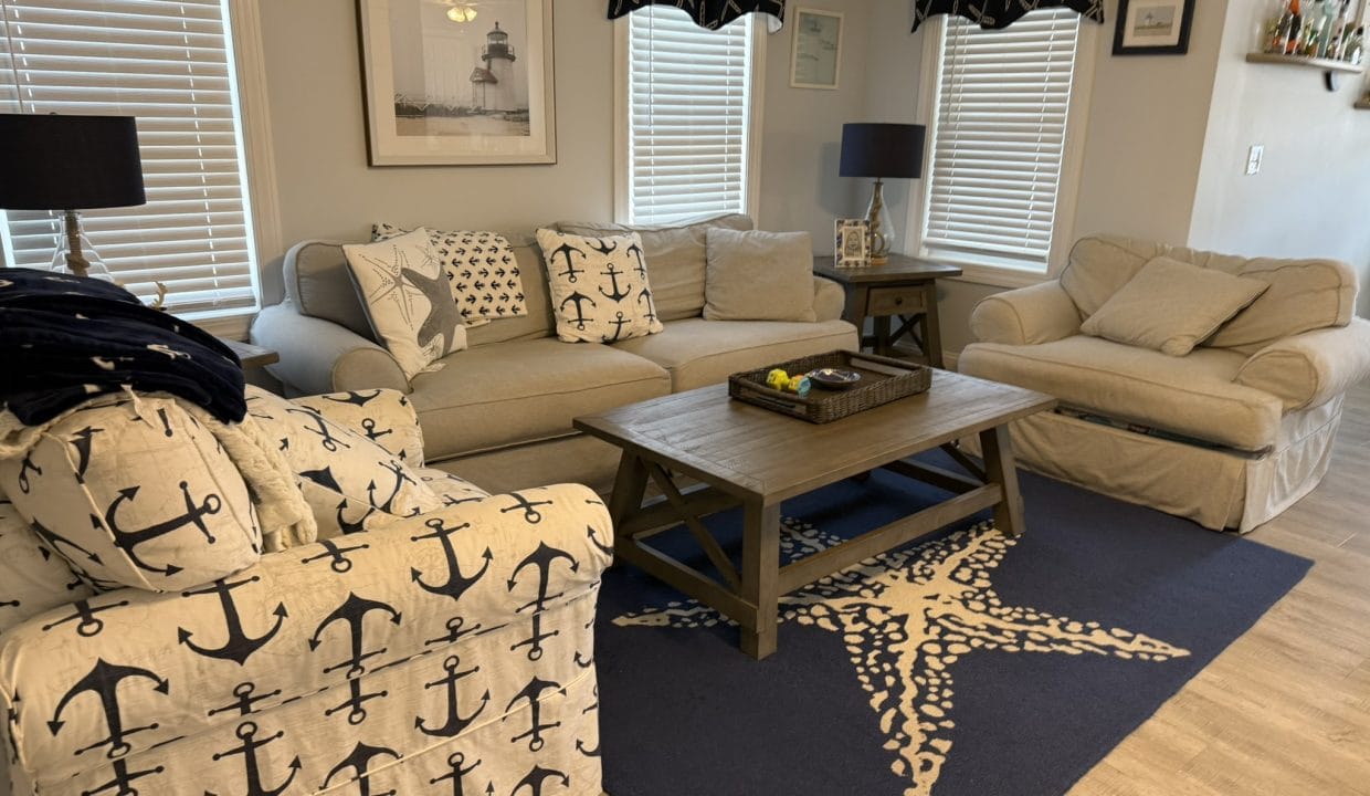 Living room with beige sofas, anchor-themed decor, navy lamps, framed lighthouse print, wooden coffee table, and starfish rug on light wood flooring.