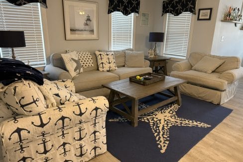 Living room with beige sofas, anchor-themed decor, navy lamps, framed lighthouse print, wooden coffee table, and starfish rug on light wood flooring.