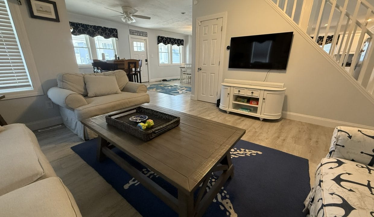 A living room with a beige sofa and chair, a wooden coffee table on a blue rug, a wall-mounted TV, and a view into a dining area and staircase.