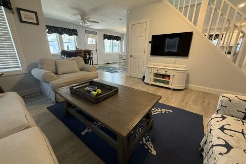 A living room with a beige sofa and chair, a wooden coffee table on a blue rug, a wall-mounted TV, and a view into a dining area and staircase.