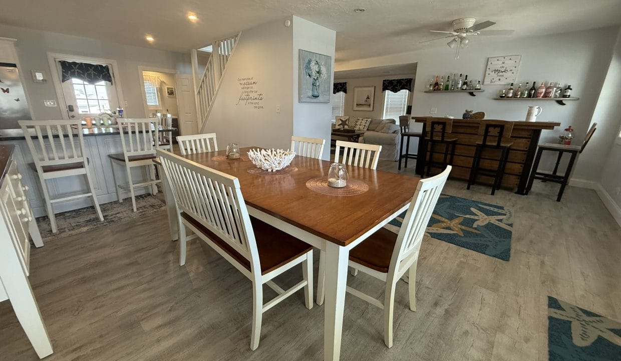 A dining room with a wooden table and six white chairs, a breakfast bar with stools, and a separate bar area in a bright, open space with light-colored floors.