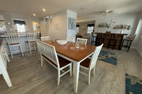 A dining room with a wooden table and six white chairs, a breakfast bar with stools, and a separate bar area in a bright, open space with light-colored floors.