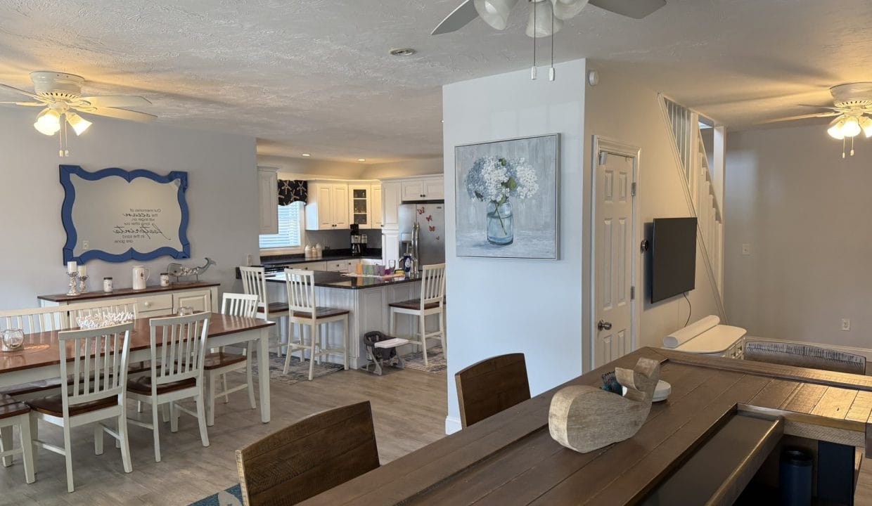 Open-concept dining and kitchen area with white chairs and wooden tables, ceiling fans, and light gray walls decorated with framed art. Stairs are visible in the background.