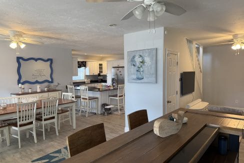 Open-concept dining and kitchen area with white chairs and wooden tables, ceiling fans, and light gray walls decorated with framed art. Stairs are visible in the background.