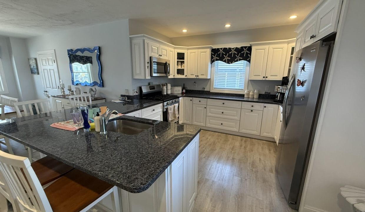 Modern kitchen with white cabinets, stainless steel appliances, black countertops, a center island with sink, and wooden stools. A window with a starfish-patterned valance is above the sink.