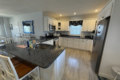 Modern kitchen with white cabinets, stainless steel appliances, black countertops, a center island with sink, and wooden stools. A window with a starfish-patterned valance is above the sink.