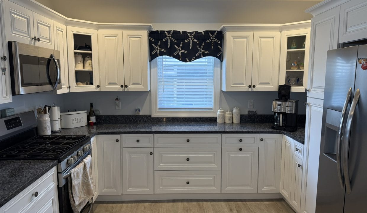 Modern kitchen with white cabinets, stainless steel appliances, black countertops, a window with a starfish-patterned valance, and light wood flooring.