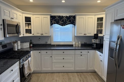 Modern kitchen with white cabinets, stainless steel appliances, black countertops, a window with a starfish-patterned valance, and light wood flooring.