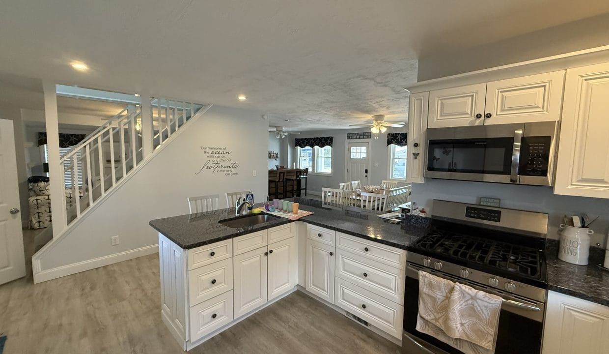 A modern kitchen with white cabinets, black countertops, a gas stove, microwave, and a view of a dining area and staircase in the background.