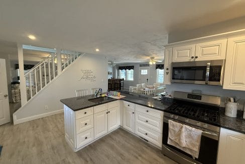 A modern kitchen with white cabinets, black countertops, a gas stove, microwave, and a view of a dining area and staircase in the background.
