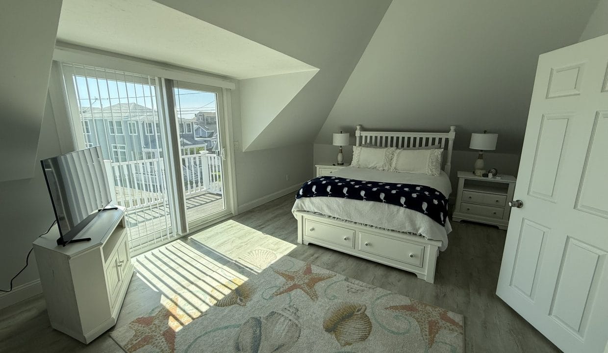 Bright bedroom with a double bed, nightstands, TV on a stand, large sliding glass doors to a balcony, and a beach-themed rug on a wooden floor.