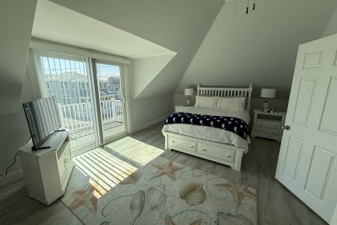 Bright bedroom with a double bed, nightstands, TV on a stand, large sliding glass doors to a balcony, and a beach-themed rug on a wooden floor.