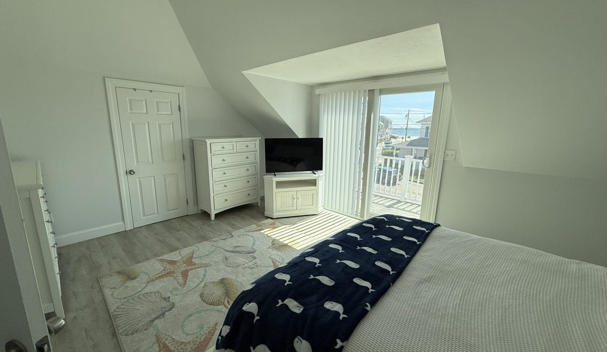 A bright bedroom with white furniture, a TV, a bed with a whale-patterned blanket, a rug with sea motifs, and sliding glass doors leading to a balcony with an ocean view.