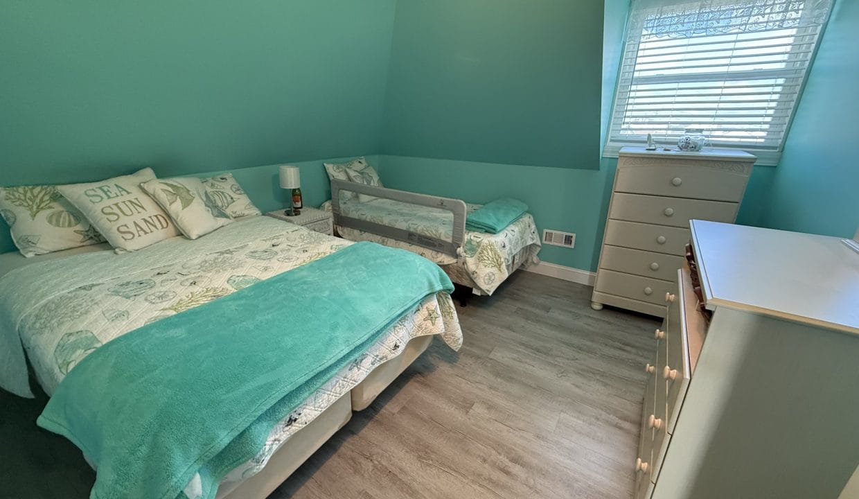 A teal-painted bedroom with a twin bed, a toddler bed with a safety rail, two dressers, and a window with blinds letting in daylight.