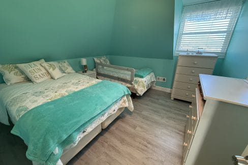 A teal-painted bedroom with a twin bed, a toddler bed with a safety rail, two dressers, and a window with blinds letting in daylight.