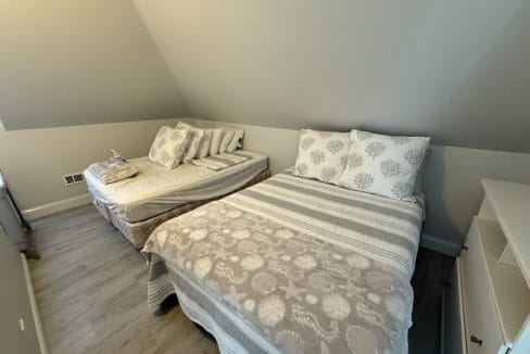 Two neatly made beds with patterned bedding are placed side by side in a small, sloped-ceiling room with hardwood floors and light-colored walls.