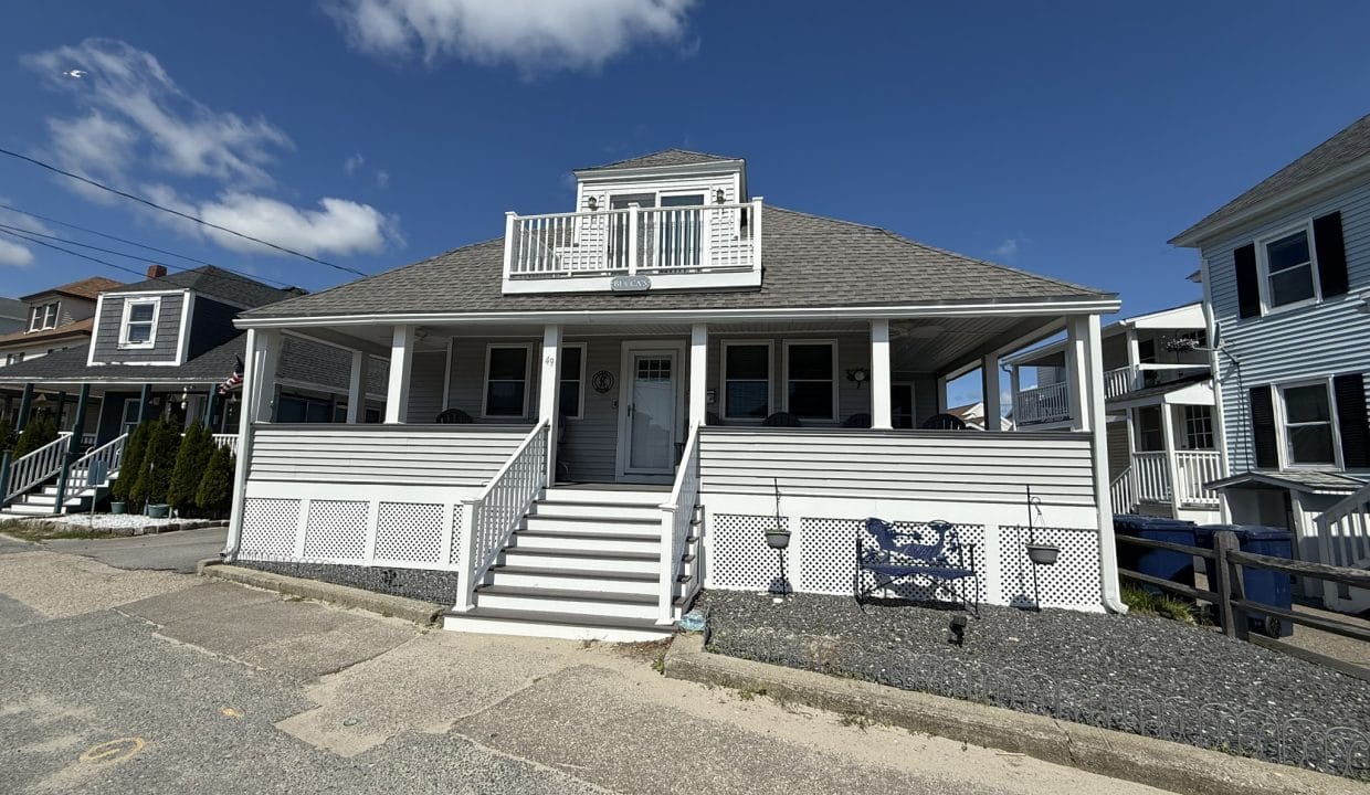 A two-story gray house with white trim, a covered wraparound porch, and steps leading to the front door, set under a blue sky with a few clouds.