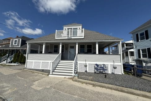 A two-story gray house with white trim, a covered wraparound porch, and steps leading to the front door, set under a blue sky with a few clouds.