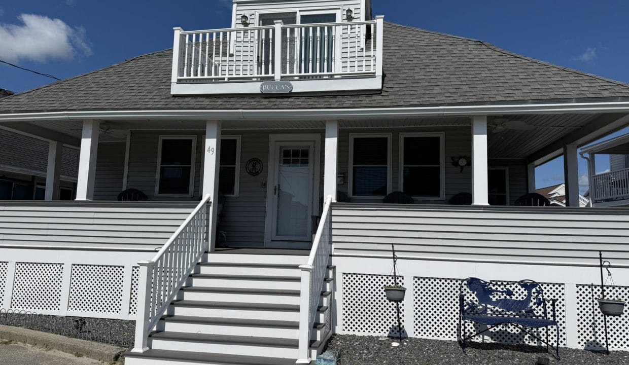 A two-story gray house with a front porch, white railings, and stairs leading up to the entrance, under a clear blue sky.