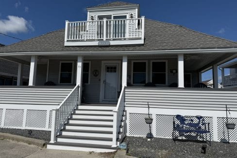 A two-story gray house with a front porch, white railings, and stairs leading up to the entrance, under a clear blue sky.