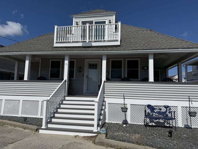 A two-story gray house with a front porch, white railings, and stairs leading up to the entrance, under a clear blue sky.
